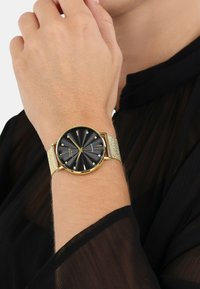 Gouden horloge met een zwarte textuur wijzerplaat en gouden accenten; mesh band; slank profiel; beschikt over duidelijke uurmarkeringen en een minimalistisch ontwerp.