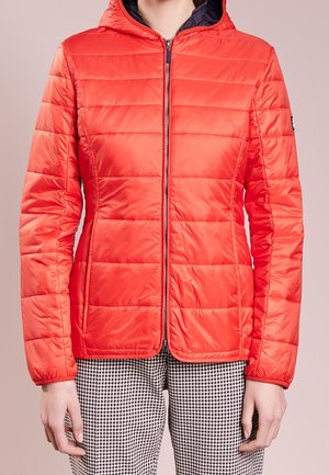 Frau trägt eine leuchtend rote, gesteppte Jacke mit Reißverschluss und Kapuze sowie schwarz-weiße Hahnentritt-Hose, steht mit entspannten Händen.