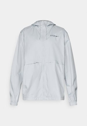Corta-vento cinza claro com fecho éclair total, capuz, punhos elásticos, cordões e pequenos logótipos "BARCA" e swoosh da Nike no lado esquerdo do peito.