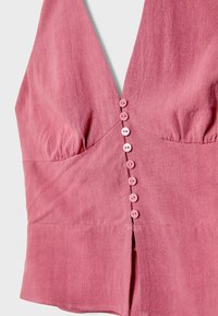 Top sans manches rose avec un décolleté en V profond, comportant huit boutons répartis uniformément sur le devant et un tissu froncé au niveau de la poitrine.