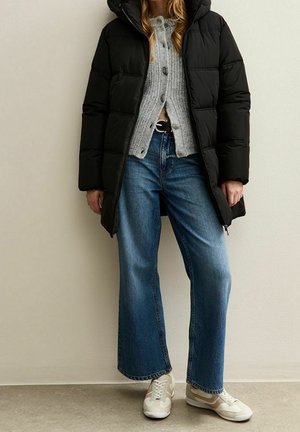 Femme portant une doudoune noire, un cardigan gris boutonné, un jean bleu à jambes larges et des baskets beiges, debout à l’intérieur contre un mur uni.