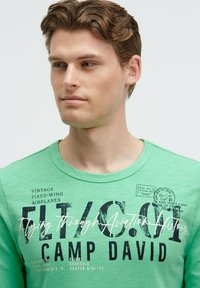 Grünes Baumwoll-T-Shirt mit Rundhalsausschnitt und in Schwarz und Weiß gedrucktem Text, im Vintage-Luftfahrtstil mit Grafiken.