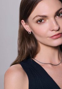 Srebrne kolczyki w kształcie obręczy z geometrycznym wzorem, noszone z ciemnym, ryflowym topem, dopełnione subtelnym naszyjnikiem łańcuszkowym. Gładka tekstura i polerowane wykończenie.