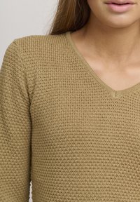 Beige knitted sweater with a V-neck design, featuring a textured pattern and ribbed edges near the neckline and cuffs.