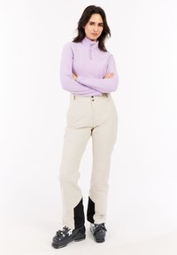 Maglia viola chiaro con zip e maniche lunghe abbinata a pantaloni crema con dettagli neri alle caviglie, indossata con stivali da sci neri.