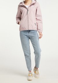 Veste rose clair en tissu doux, avec une capuche, une fermeture éclair à l'avant et deux poches latérales. Portée sur un jean bleu clair et des baskets.