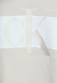 Camiseta de color beige claro con una banda horizontal blanca que presenta el logo "CK" en relieve en plata pálido. Tejido de mezcla de algodón suave con textura lisa.