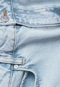 Hellblaues Denimgewebe mit einem strukturierten Muster, das orangefarbene Nähte und metallene Knopfdetails aufweist. Nahaufnahme des Materials und des Designs.