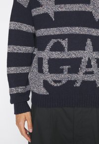 Pull en maille bleu foncé avec des rayures horizontales grises et de grandes lettres texturées partielles sur la poitrine et les manches.