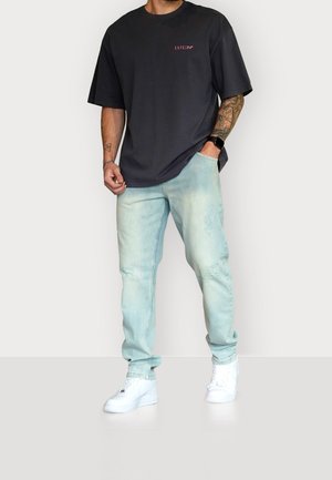 Mann trägt ein schwarzes oversized T-Shirt mit dem Schriftzug "Esteem", hellblaue Destroyed-Jeans, weiße Sneakers und eine Smartwatch, steht mit einer Hand in der Tasche.