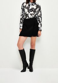 Černá mini sukně s třásněmi v kombinaci s černobílou vzorovanou košilí s dlouhým rukávem. Modelka má na sobě černé kozačky na vysokém podpatku sahající až ke stehnům.
