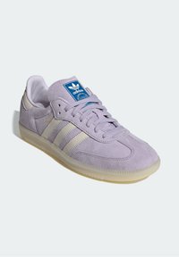 Zapatillas de ante moradas con tres rayas blancas, punta redonda y suela de goma. Presenta un parche de logo azul en la lengüeta.