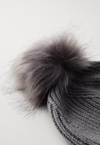 Bonnet en tricot gris avec un grand pompon en fausse fourrure grise, doux au toucher, sur le dessus. Le chapeau présente un design texturé et un revers roulé.