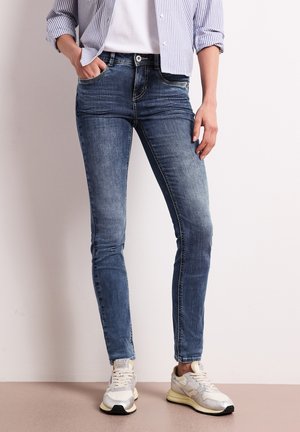 Kvinde iført falmede blå skinny jeans, hvide sneakers og en stribet langærmet skjorte delvist stoppet ind med den ene hånd i lommen.