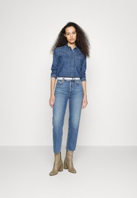 Camisa de gangue com mangas longas, bolso frontal; jeans azuis com silhueta ajustada; botas de salto alto até ao tornozelo em cinza claro; cinto branco com fivelas circulares.