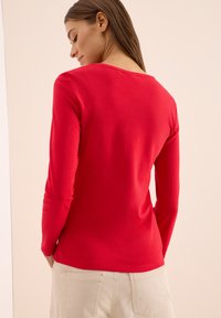 Langärmliges rotes Oberteil aus Baumwolle mit rundem Ausschnitt, das eine glatte Textur und eine figurbetonte Silhouette aufweist, getragen mit hellen Hosen.
