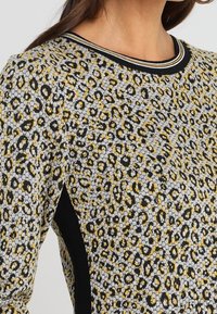 Top en tricot à imprimé léopard avec un motif gris, jaune et noir. Présente un col côtelé noir et des accents de panneaux latéraux.