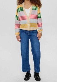 Osoba oblečená v barevném pruhovaném kardiganu, bílém tričku, modrých džínách a černých mokasínech, stojící na jednobarevném pozadí.