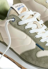 Zapatillas con parte superior de ante beige y verde, cordones blancos, acentos de malla y una etiqueta con un diseño de olas en azul y dorado en la lengüeta.