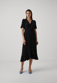 Black wrap dress with short puff sleeves, a flared hem, and a tie at the waist. Smooth fabric with a subtle texture. Paired with light blue heels.