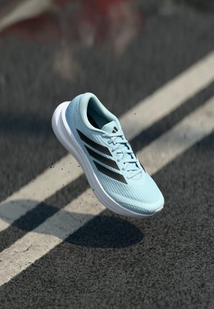 Scarpa da corsa azzurro chiaro con strisce nere, suola bianca e lacci, sospesa in aria sopra una strada asfaltata con segnaletica bianca.