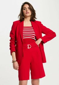 Blazer rosso con spalline, pantaloni corti rossi coordinati e una maglietta a righe rossa e bianca. Presenta una cintura con un dettaglio della fibbia dorata.