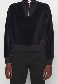 Pullover in velluto nero con collo a imbuto, zip argentata e vestibilità ampia. I polsini e l'orlo a coste aggiungono texture. Abbinato a pantaloni neri.