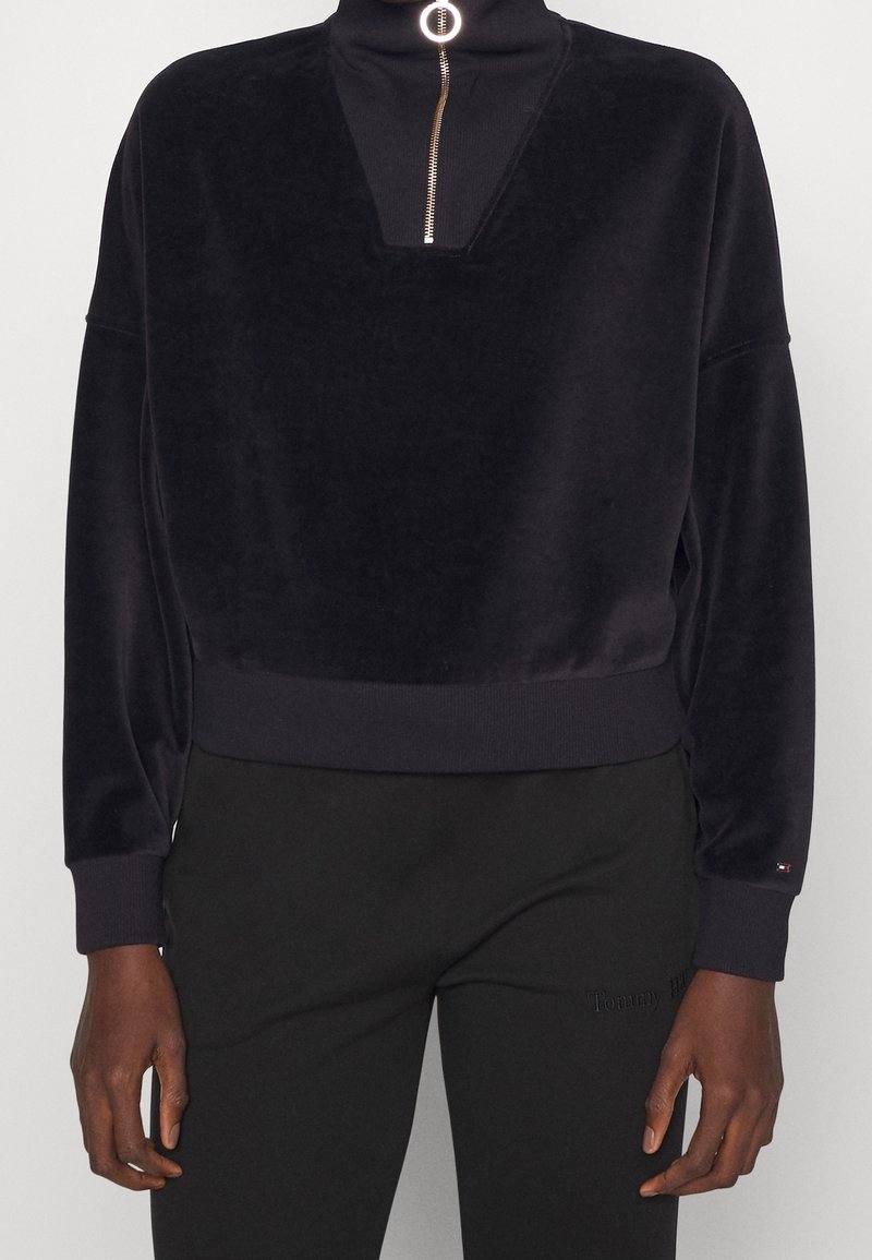 Pullover in velluto nero con collo a imbuto, zip argentata e vestibilità ampia. I polsini e l'orlo a coste aggiungono texture. Abbinato a pantaloni neri.