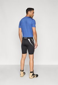 Camisola atlética azul de manga curta com padrão texturizado, combinada com calções de compressão pretos com detalhes cinzentos e logótipo, e botas claras.