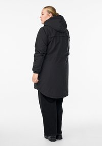 Zwarte lange coat met capuchon, van een soepele stof, met elastische manchetten en een gebogen zoom. Draag dit over zwarte broek met een minimalistisch ontwerp.
