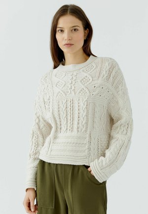 Vrouw met lang bruin haar, die een gebreide off-white trui met patroon en olijfgroene broek draagt, met een hand in haar zak, tegen een effen achtergrond.