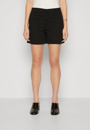 Frau trägt schwarze knielange Shorts, schwarze Schuhe mit Absatz und ein weißes Oberteil und steht vor einem schlichten weißen Hintergrund.