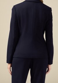 Blazer bleu marine à coupe ajustée, dotée d'un col cranté et d'une coupe droite. Tissu lisse avec une légère brillance, souligné par des coutures minimales.