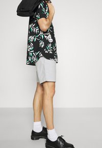 Camisa negra de flores con mangas cortas y hojas verdes y blancas, pantalones cortos gris claro, y zapatos negros con calcetas blancas.