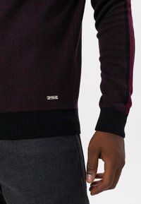 Pull en maille de couleur bordeaux foncé avec tissu texturé. Comprend des poignets et un ourlet côtelés bleu marine, ainsi qu'une petite étiquette logo en métal sur le bas devant.