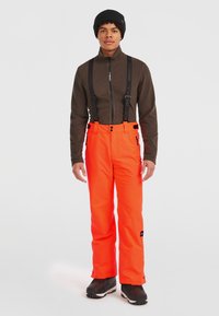 Klart orange skidbyxor med justerbara svarta hängslen, rak skärning och sidodragkedjor, i kombination med en brun zip-up jacka och svart mössa.