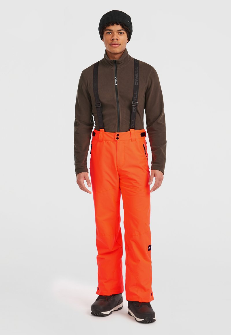 Klart orange skidbyxor med justerbara svarta hängslen, rak skärning och sidodragkedjor, i kombination med en brun zip-up jacka och svart mössa.
