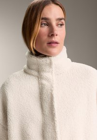 Giacca in pile color crema con colletto alto, texture morbida e dettagli di cuciture sottili. Il design è privo di parti metalliche o decorazioni visibili.