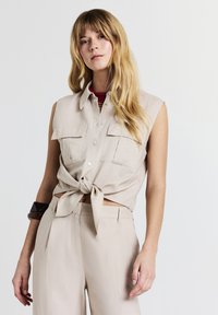 Donna con lunghi capelli biondi che indossa una camicia beige senza maniche con bottoni legata in vita, pantaloni beige e un braccialetto scuro sul braccio sinistro.