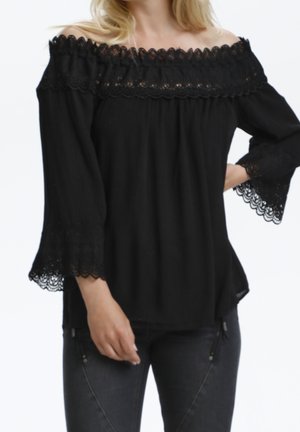 Kobieta w czarnej bluzce z odkrytymi ramionami z koronkowym wykończeniem przy dekolcie i rękawach, w ciemnych dżinsach, na białym tle.