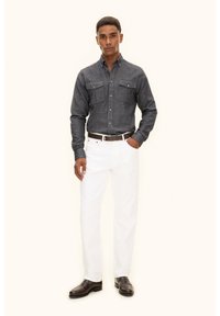 Camicia grigia con bottoni e due tasche sul petto, pantaloni bianchi e cintura marrone. Il modello indossa scarpe scure, in piedi contro uno sfondo neutro.