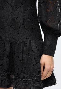 Black lace dress featuring a floral pattern, ruffled hem, and sheer balloon sleeves with textured detailing. Smooth fabric texture overall.