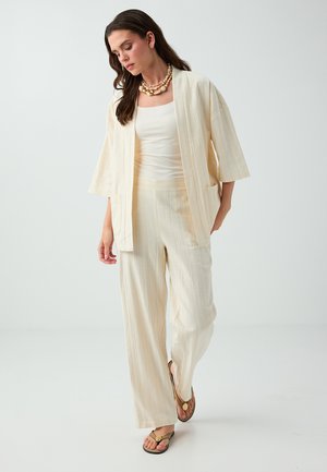 Femme portant un pantalon ample couleur crème, une veste ouverte assortie, un débardeur blanc, des tongs et un collier superposé de coquillages, marchant sur fond blanc.