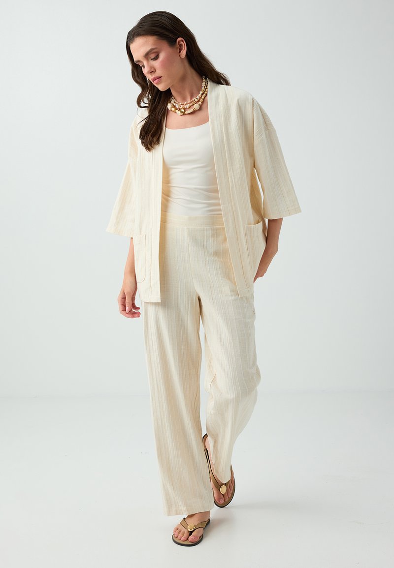 Femme portant un pantalon ample couleur crème, une veste ouverte assortie, un débardeur blanc, des tongs et un collier superposé de coquillages, marchant sur fond blanc.