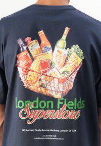Person som har på seg en mørk skjorte med en fargerik illustrasjon av en handlekurv og teksten "London Fields Superstore" på baksiden.