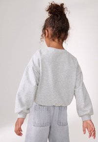 Szürke pulóver ejtett vállal, puha tapintással és bordás mandzsettákkal, világosszürke farmernadrággal párosítva, melynek két hátsó zsebe van.