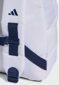 Bijela ruksak s tamnoplavim trakama i hardverom, sa Adidas logotipom u tamnoplavoj boji. Materijal je teksturiran, s urednim, strukturiranim dizajnom.