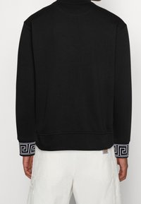 Felpa nera con maniche lunghe, colletto alto e polsini decorati con un design geometrico. Realizzata in un tessuto testurizzato.