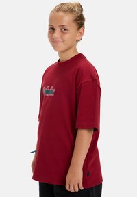 Rode katoenen t-shirt met korte mouwen, een ronde hals en zijsplitten. Bevat geborduurde tekst in wit en blauw over de borst.
