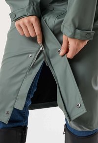 Hænder, der lukker lynlås og trykknapper på en grøn vandtæt jakke, der bæres over blå jeans og sorte støvler.